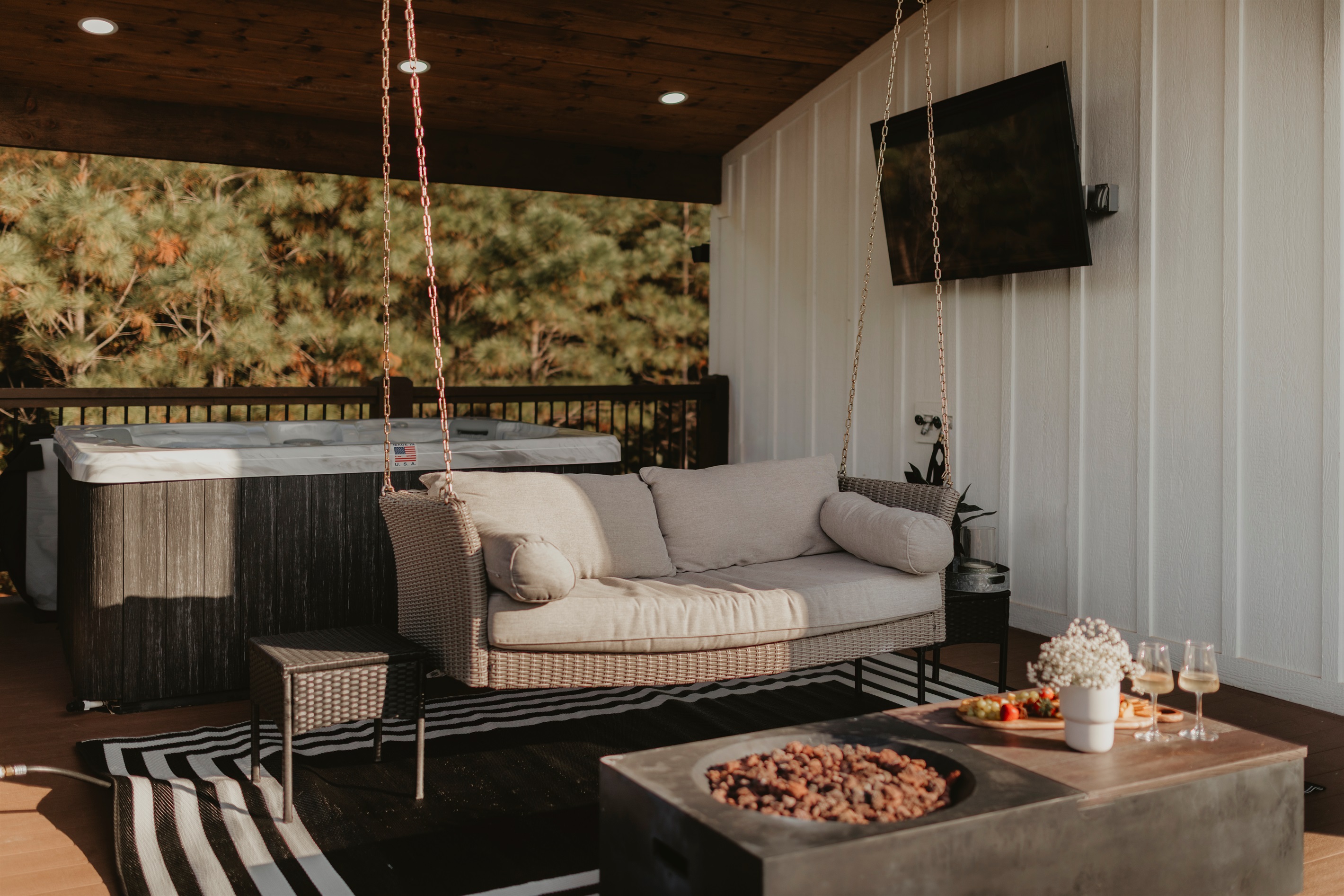  Covered patio on the second deck with a swinging sofa, fire table, hot tub, and outdoor TV. The perfect spot to unwind, watch a show, or sip drinks by the fire while surrounded by peaceful forest views.