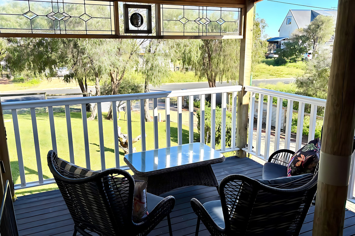 The front porch at Lake Views & Coastal Escape has leadlight panels overhead, a glass-top table and cushioned chairs, looking over the front lawn and gardens to the quiet street.