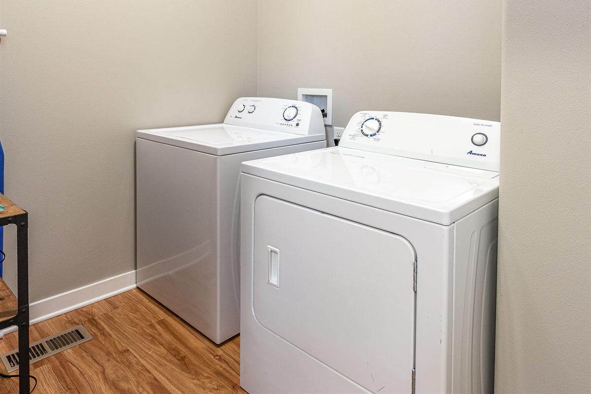 A white washing machine and dryer are side by side in a small laundry room with light-colored walls and wooden flooring.