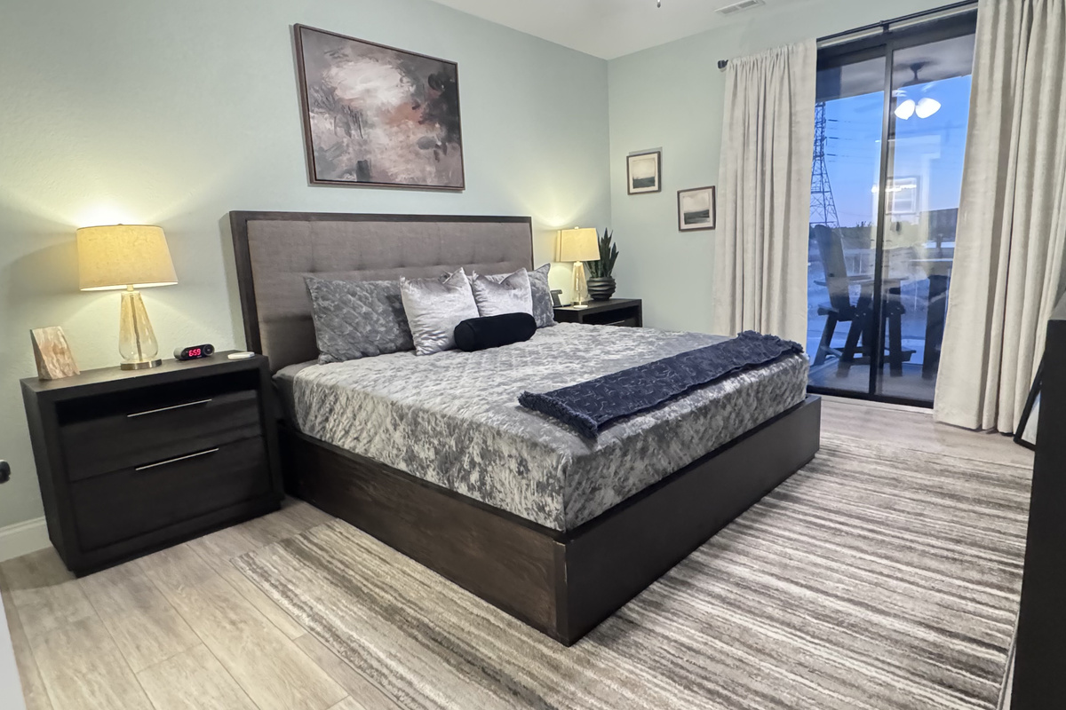 Primary Bedroom with a lakeside view and balcony access. Includes a Smart TV, large dresser, full-length mirror, two nightstands, table lamps, all bed linens, extra pillows, blanket, and cell phone/watch charging stations.