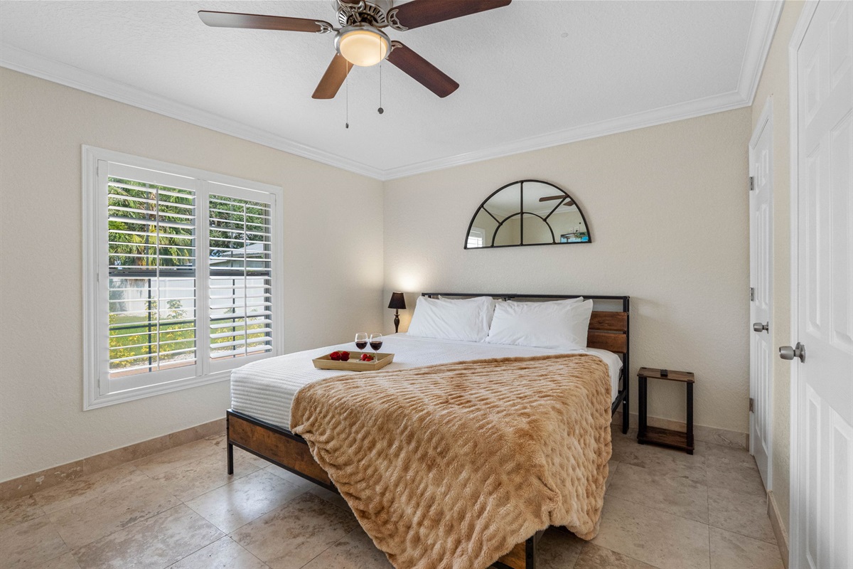 This inviting bedroom features a warm, textured throw, a rustic wood-and-metal bed frame, and plenty of natural light through plantation shutters.