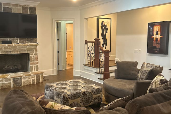 Finished basement living area featuring a stone fireplace with wall-mounted TV, recessed lighting, and crown molding. Spacious seating area flows into an open layout with staircase detail, ideal for relaxing or entertaining.