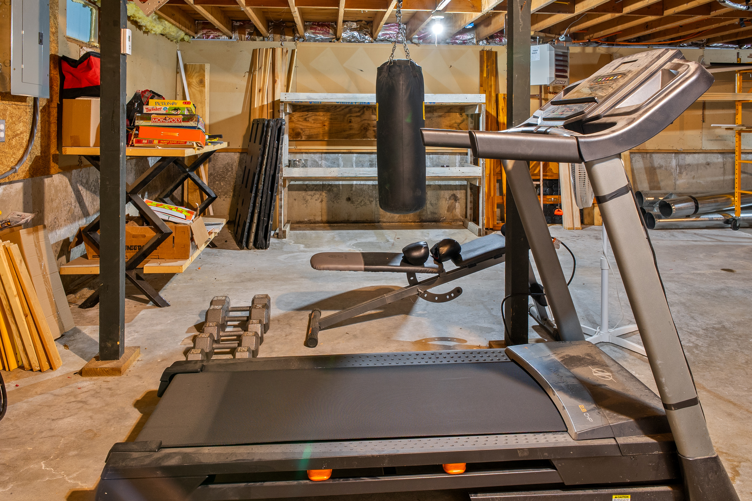 Log some miles on the motorized treadmill, positioned directly inside the basement fitness area. This alternate angle highlights the workout bench, hanging punching bag, and a shelving unit stocked with popular board games for family fun.