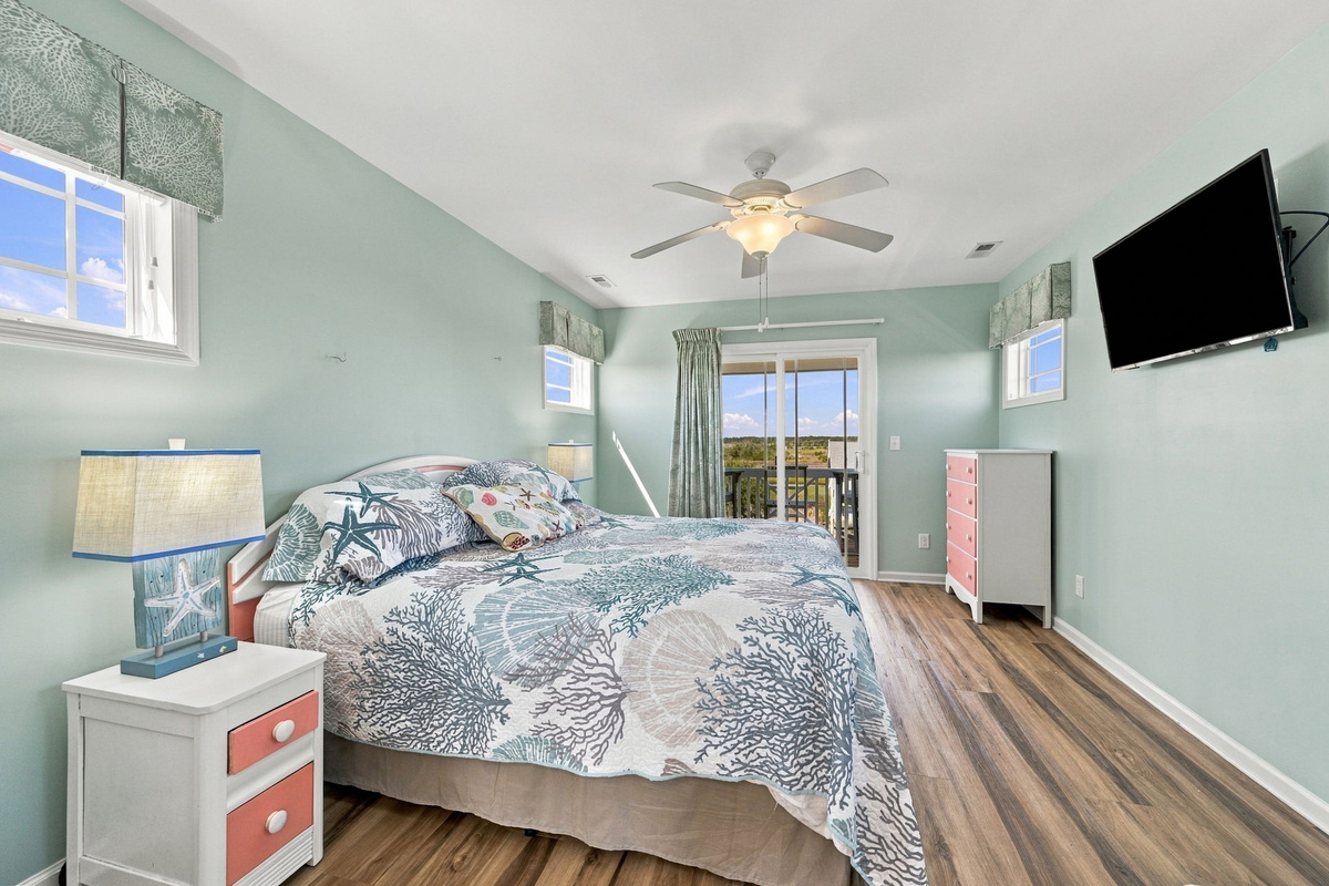 Comfortable coastal-inspired bedroom featuring a spacious bed, wall-mounted TV, and plenty of natural light