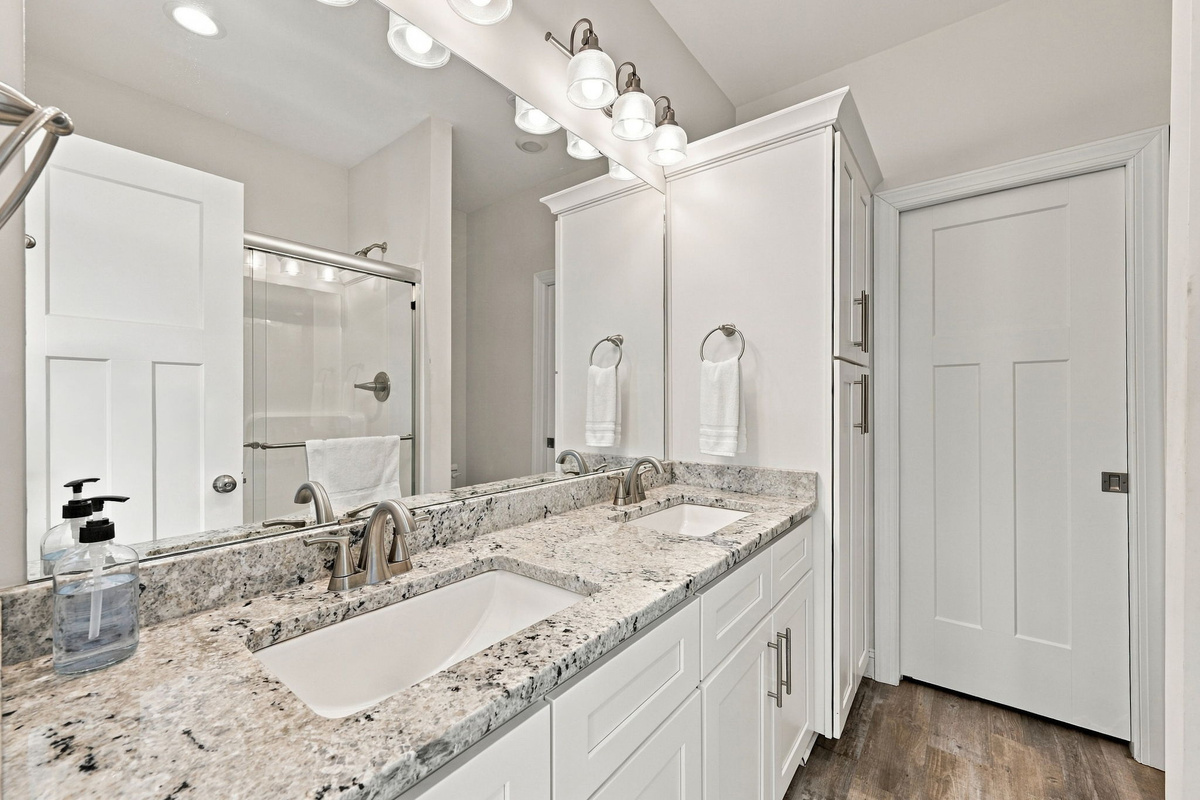 Full bathroom featuring a white double-sink vanity with granite countertops, custom cabinetry, a large mirror, and a glass-enclosed tub/shower combo.  