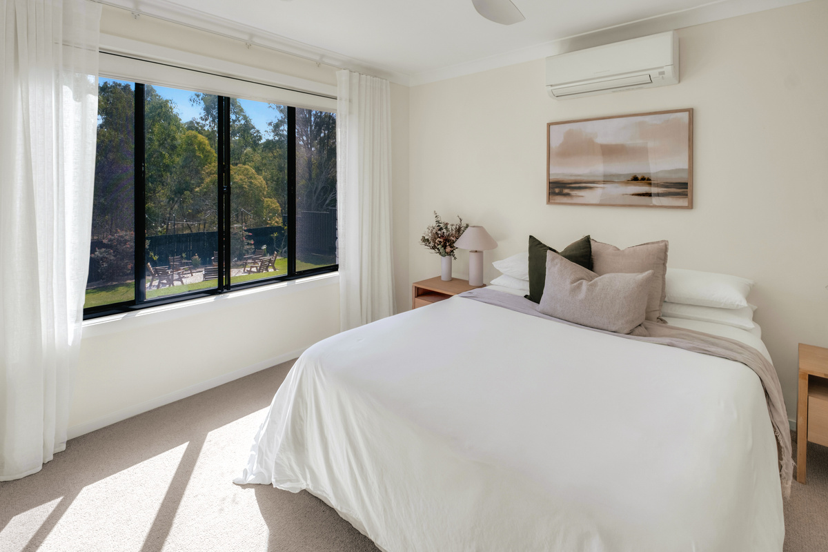 Bright and peaceful bedroom with soft neutral styling, air conditioning and large windows overlooking the leafy outdoor setting.