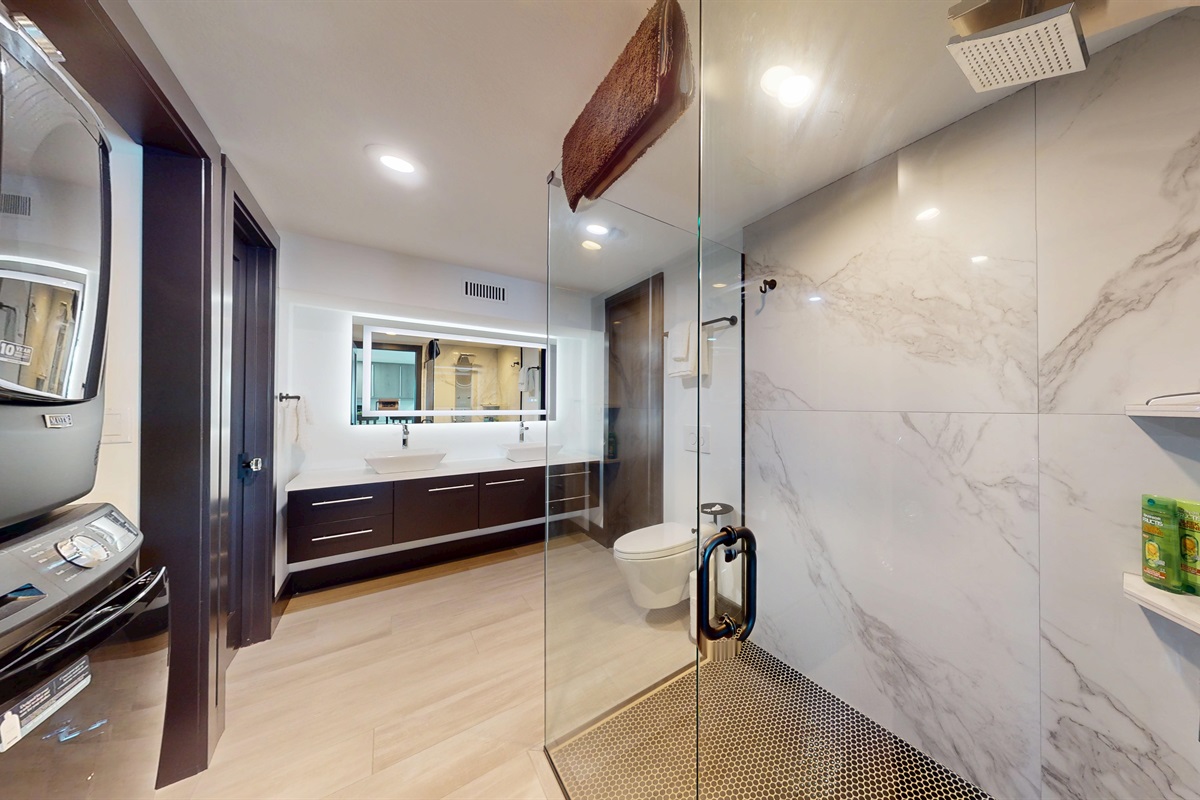 Lower-level full bathroom with double sinks, a large all-glass walk-in shower with a full-body sprayer and rainhead, plus a stackable washer and dryer.