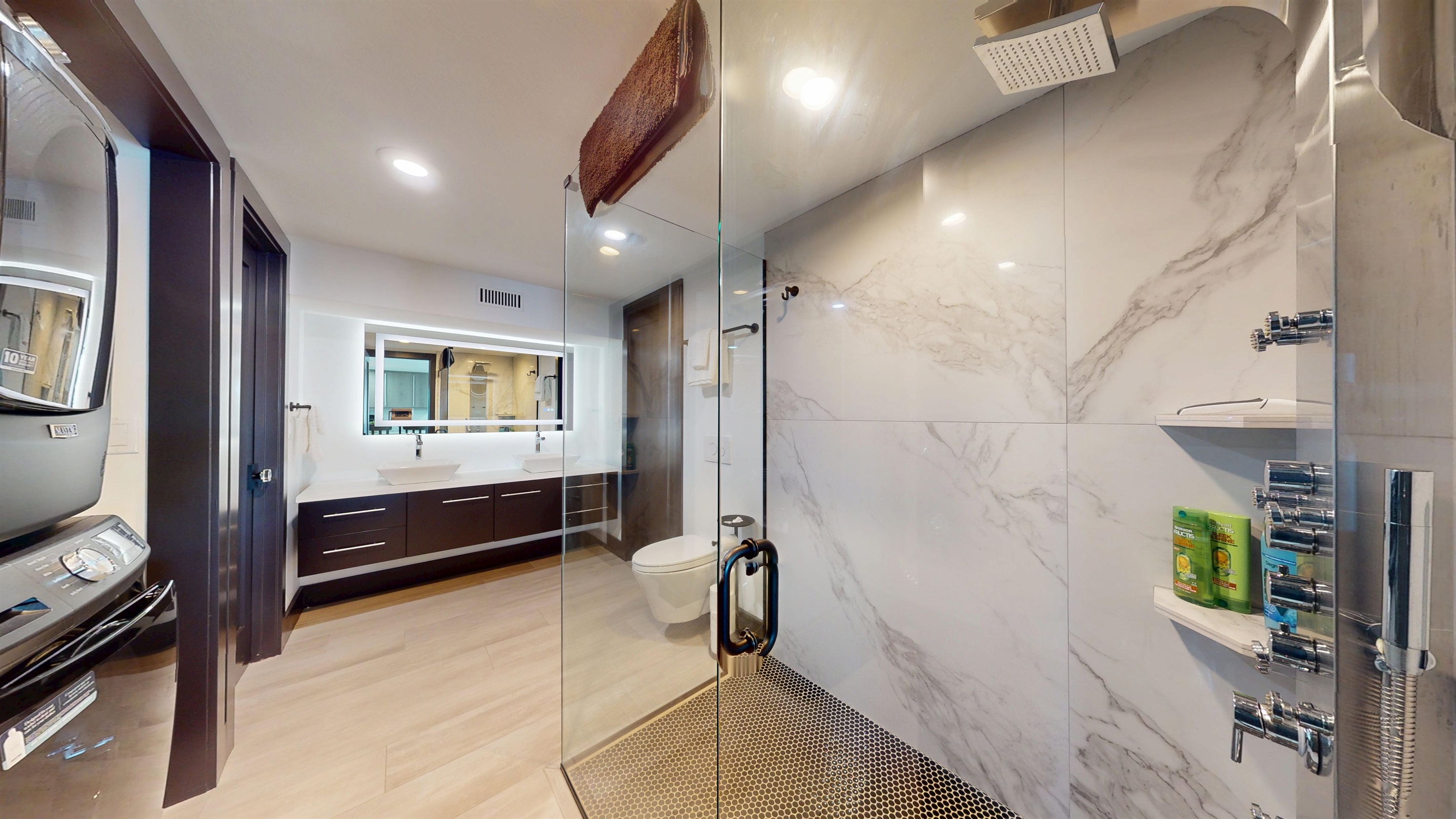 Lower-level full bathroom with double sinks, a large all-glass walk-in shower with a full-body sprayer and rainhead, plus a stackable washer and dryer.