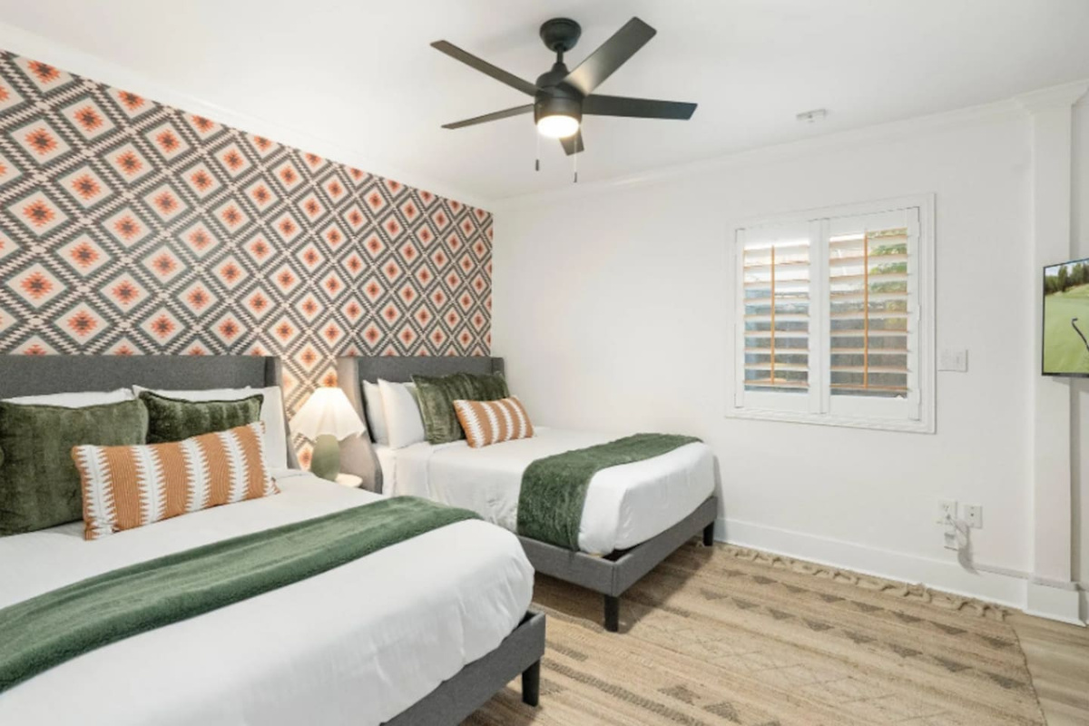 Two double beds sit beneath a bold geometric print, with layered olive and rust pillows tying the room together. Soft natural light comes through the shutters, making this a bright, comfortable spot for friends or family sharing a room.

