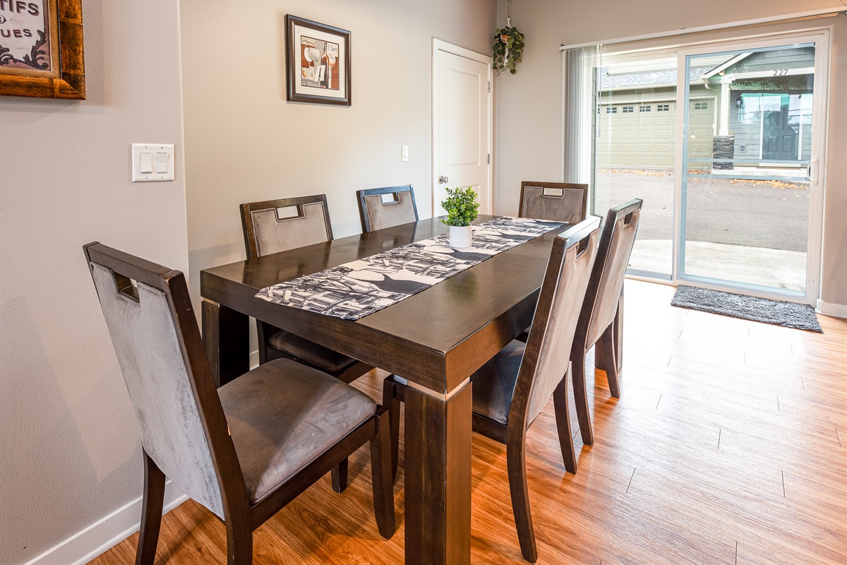 A modern dining room features a dark wooden table with a patterned runner, surrounded by six upholstered chairs, and a small green plant in the center, with a sliding glass door leading to the outside visible in the background.