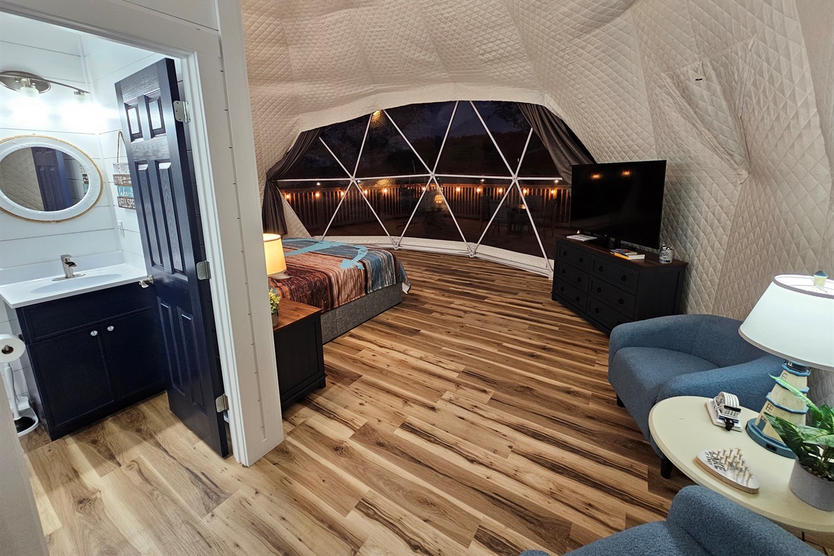 Cozy nighttime interior with king bed, lounge seating, TV, and private bathroom — warm, inviting, and steps from the glowing deck