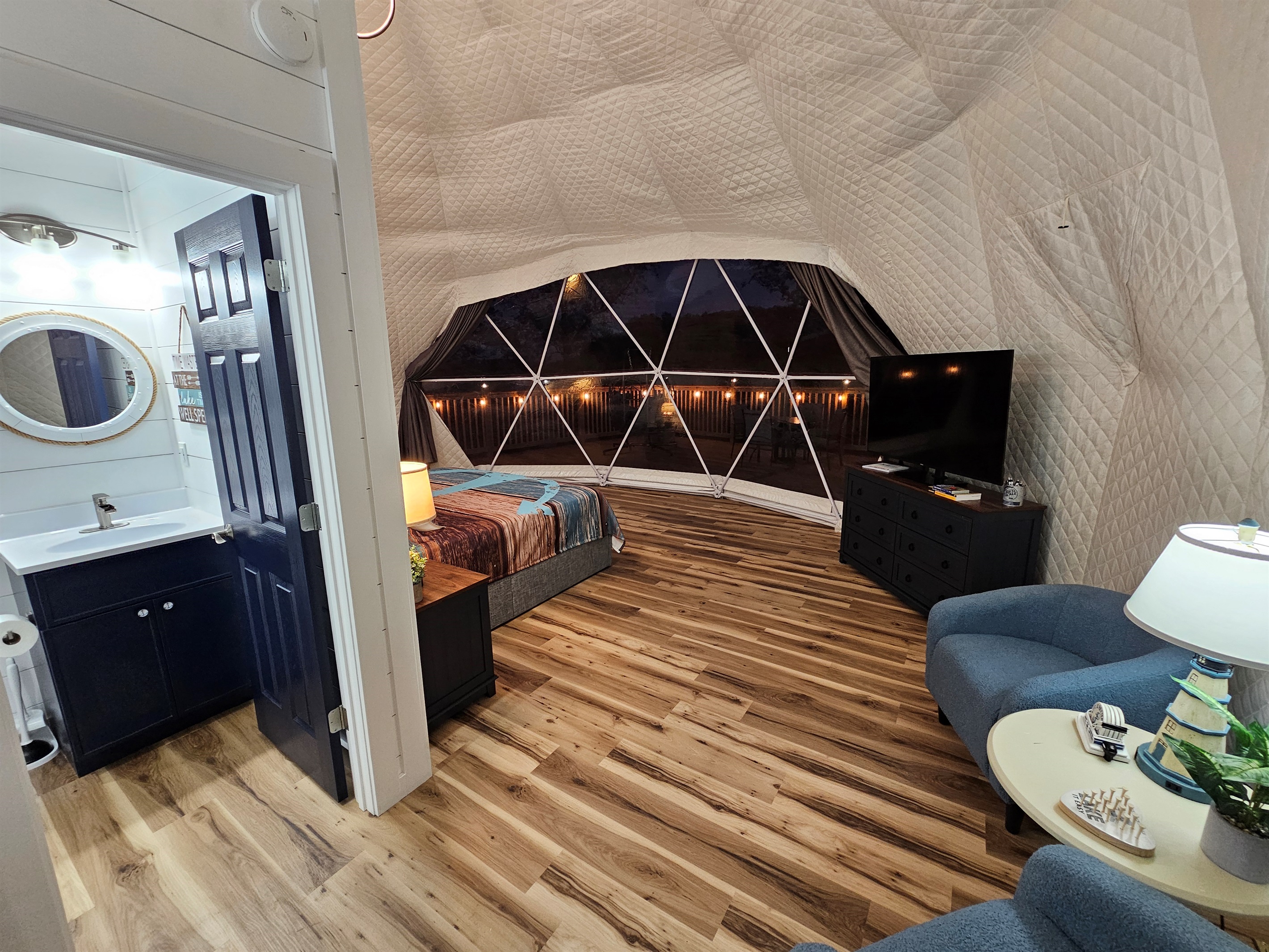 Cozy nighttime interior with king bed, lounge seating, TV, and private bathroom — warm, inviting, and steps from the glowing deck