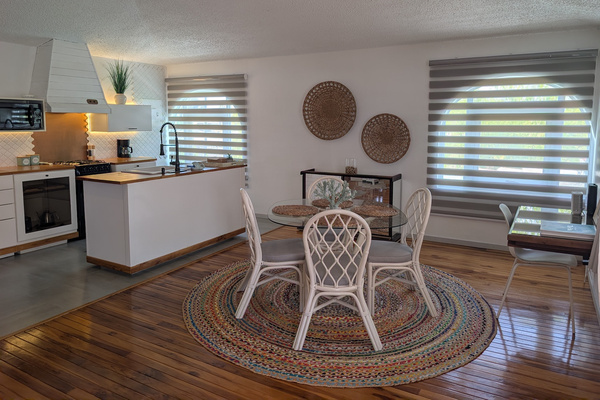 Open-concept upstairs kitchenette and dining area at Villa Coral featuring a round glass table with white chairs, a woven jute rug, hardwood floors, and modern white cabinetry in Dorado, Puerto Rico.
