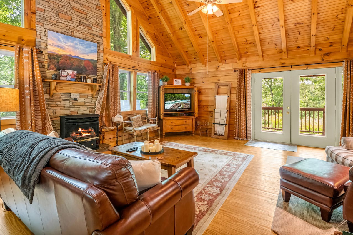Main living area featuring high vaulted wooden ceilings with exposed beams, stone fireplace with a mantel, french doors opening to the back deck, television set, leather sofa and ottoman, and a wooden coffee table