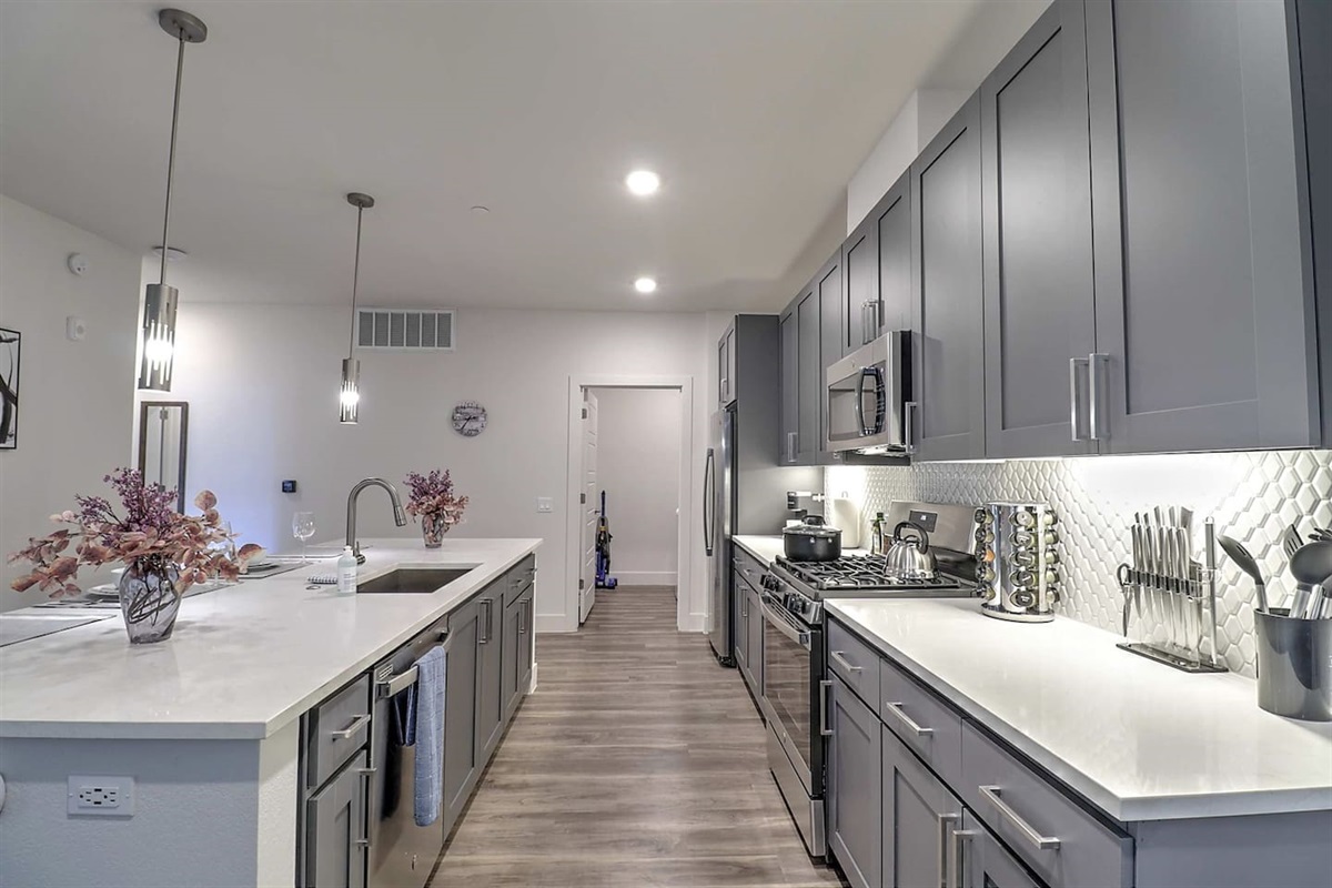 - Step into a spacious kitchen with elegant gray cabinets and sleek countertops   - Bright lighting enhances the open layout and stainless steel appliances   - Ideal for cooking meals, entertaining, or enjoying casual morning coffee