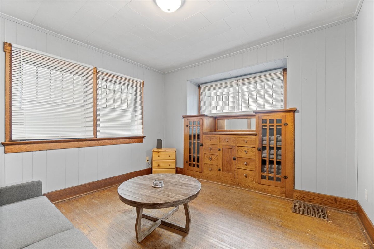 Charming living room featuring a cozy bed, stylish decorations, large windows, a tiny table in the middle, a drawer in the corner, and a cabinet—accentuated by sleek wood flooring, creating a comfortable and functional space with modern appeal