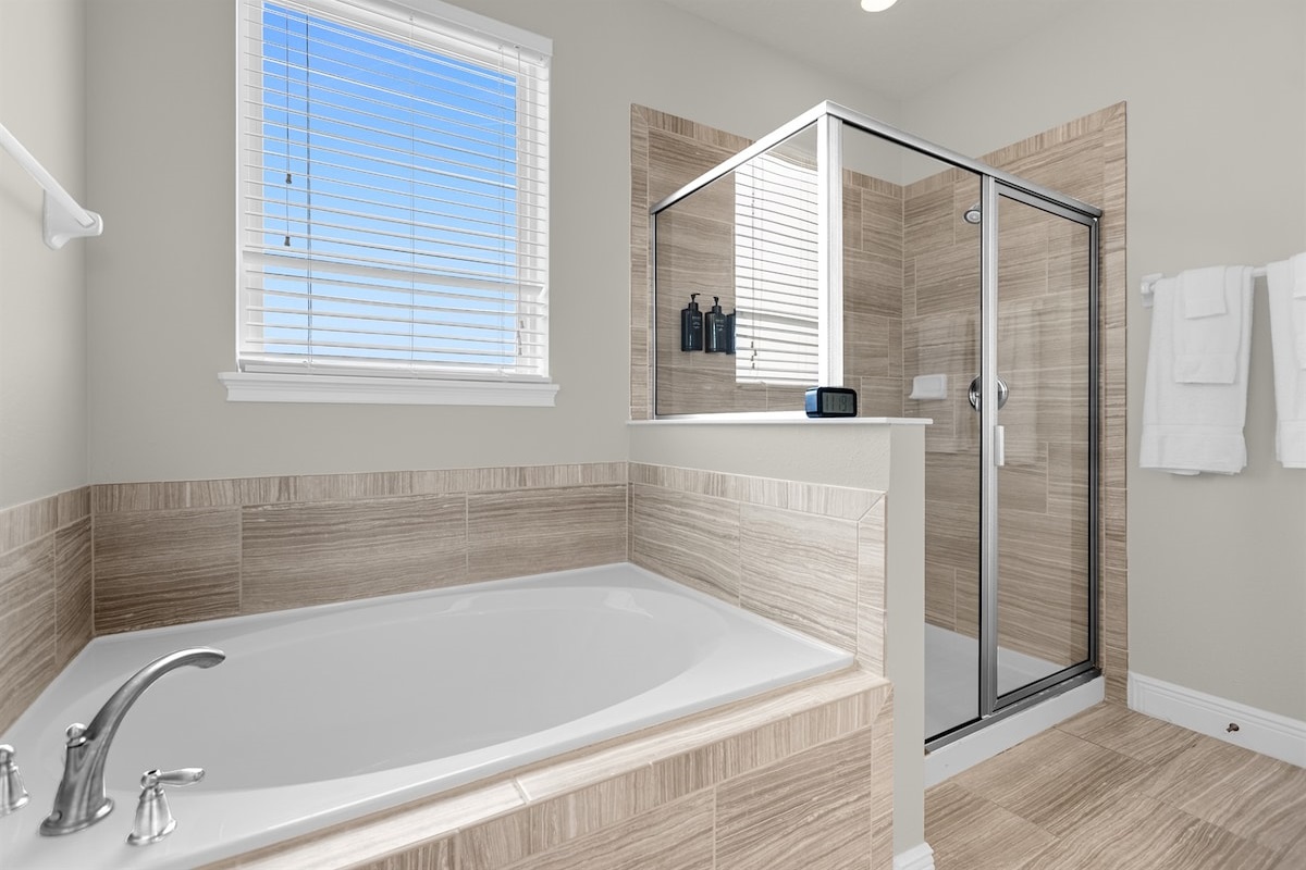 Bright bathroom featuring a glass-enclosed shower beside a spacious bathtub, with a window above the tub allowing natural light to fill the space. The clean design and airy feel create a relaxing, spa-like atmosphere for your stay.