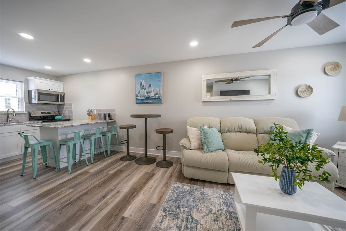 Spacious open living and dining area with comfortable seating, ceiling fan, and soft coastal decor. The layout connects seamlessly to the kitchen and bar seating, creating an easygoing space for relaxing, dining, and spending time together.