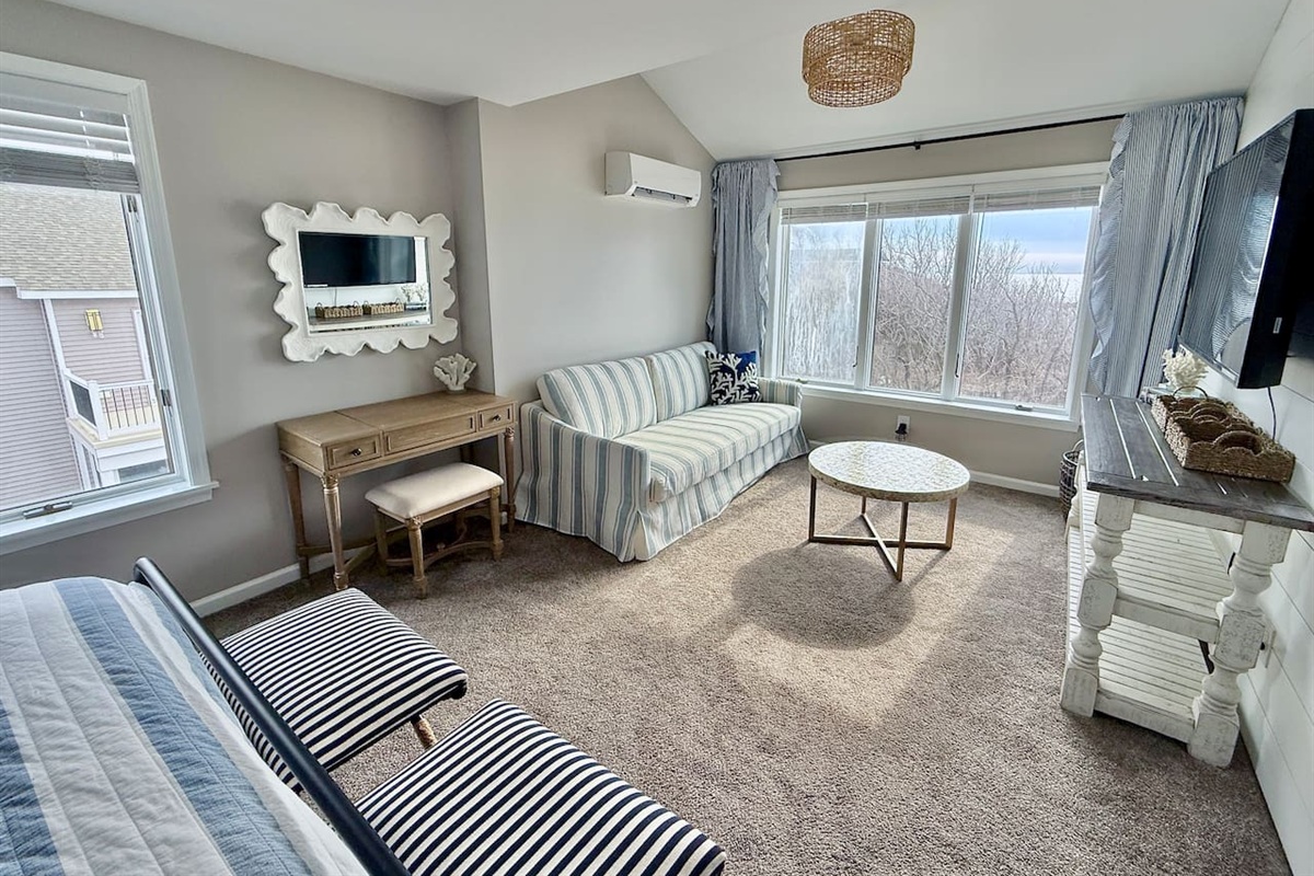 The master bedroom features this bright, versatile living space features a cozy striped sofa, a wooden vanity with a decorative mirror, and scenic windows, creating a perfect, light-filled retreat for guests to relax during their stay.
