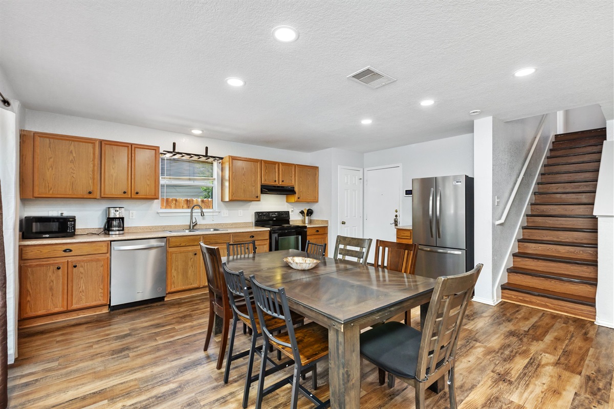 Open-concept main floor with kitchen, dining table, stainless refrigerator, and living room all connected. Hardwood floors and staircase leading to 4 upstairs bedrooms. Northeast San Antonio group rental, sleeps 10.