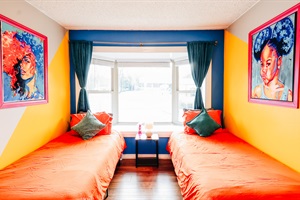 Refreshing Retreat: Twin Beds in Zesty Orange, Set Against Serene Bluish-Green Curtains, Harmonize with the Soft Green, Yellow, and White Tones of the Walls in this Tranquil Bedroom. Enjoy Natural Light Through the Window and Entertainment on the TV Screen
