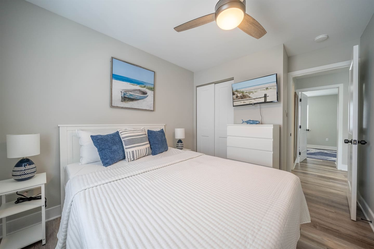 Calm, coastal inspired bedroom with a comfortable bed, soft linens, and ceiling fan for restful sleep. Thoughtful decor and a wall mounted TV create a relaxing retreat after a full day at the beach or exploring the area.
