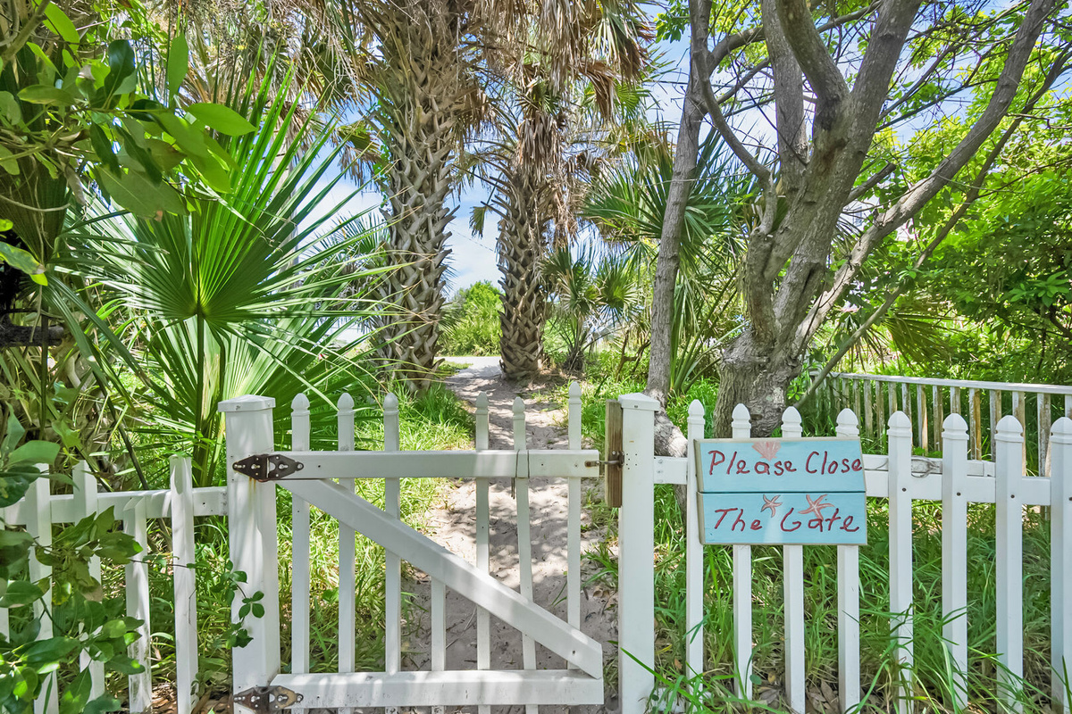 The white picket gate onto the beach path, kept closed to protect the dunes behind it. Please latch it after you; the sea oats hold this whole coastline together and they need all the help they can get.