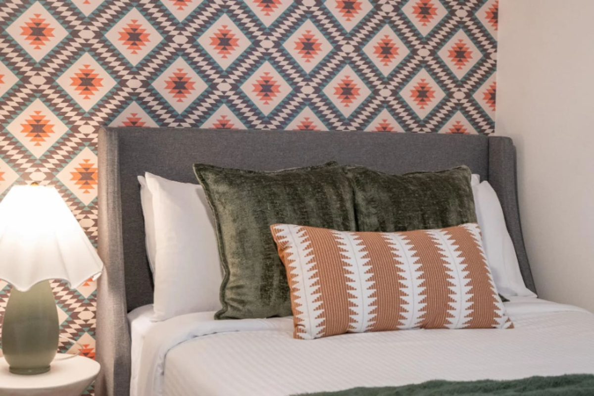 A bold Southwest-print wall backs a plush upholstered headboard, layered with olive velvet and rust-striped pillows for a cozy, textured finish.
