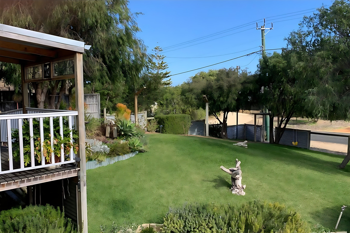 The fully fenced front lawn has garden beds, a driftwood feature and shady trees, seen from the porch looking across the grass towards the quiet street at Preston Beach.