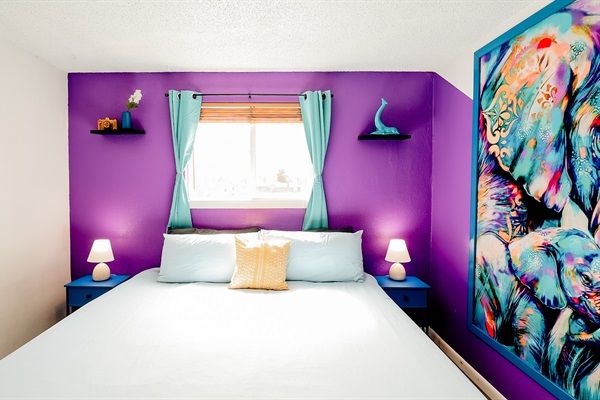 A soft purple wall whispers tranquility, setting a peaceful backdrop for a crisp white bed and a glowing lamp. It's a haven for restful nights and dreamy mornings, where comfort meets simplicity in every detail.