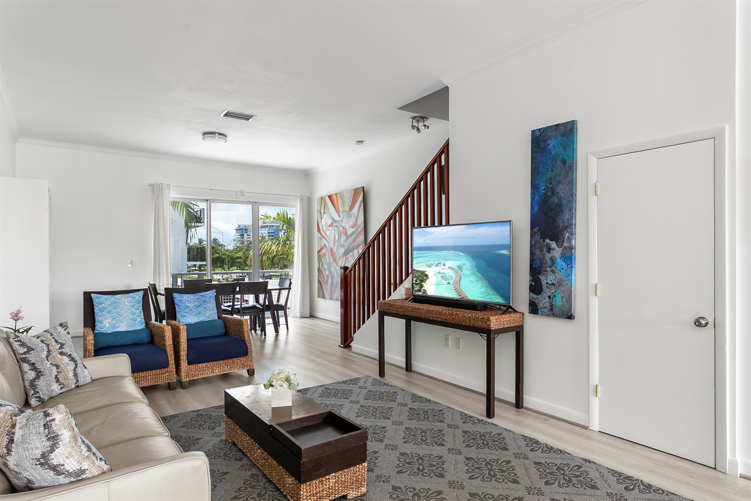 Living room with leather sofa, accent chairs, and a 50-inch flat screen TV. Sliding glass doors open to a Juliet balcony for sea breeze.