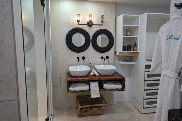 Symmetrical view of the primary bathroom at Villa Coral displaying a custom live-edge wood vanity, vessel sinks, a glass steam shower, built-in closet organization, and travertine tile walls in Dorado, Puerto Rico.
