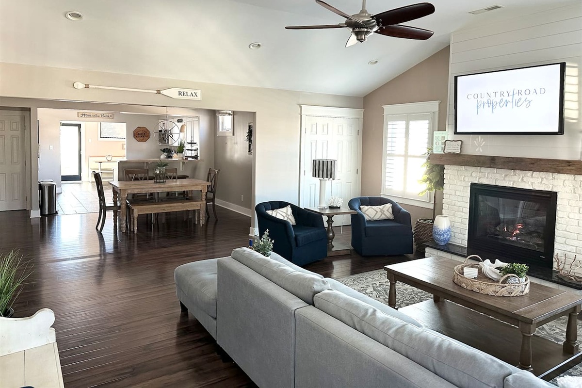 This open-concept layout connects the living and dining areas, featuring vaulted ceilings and a cozy fireplace. The space includes a large sectional and a farmhouse-style dining table, perfect for family meals or a movie night.