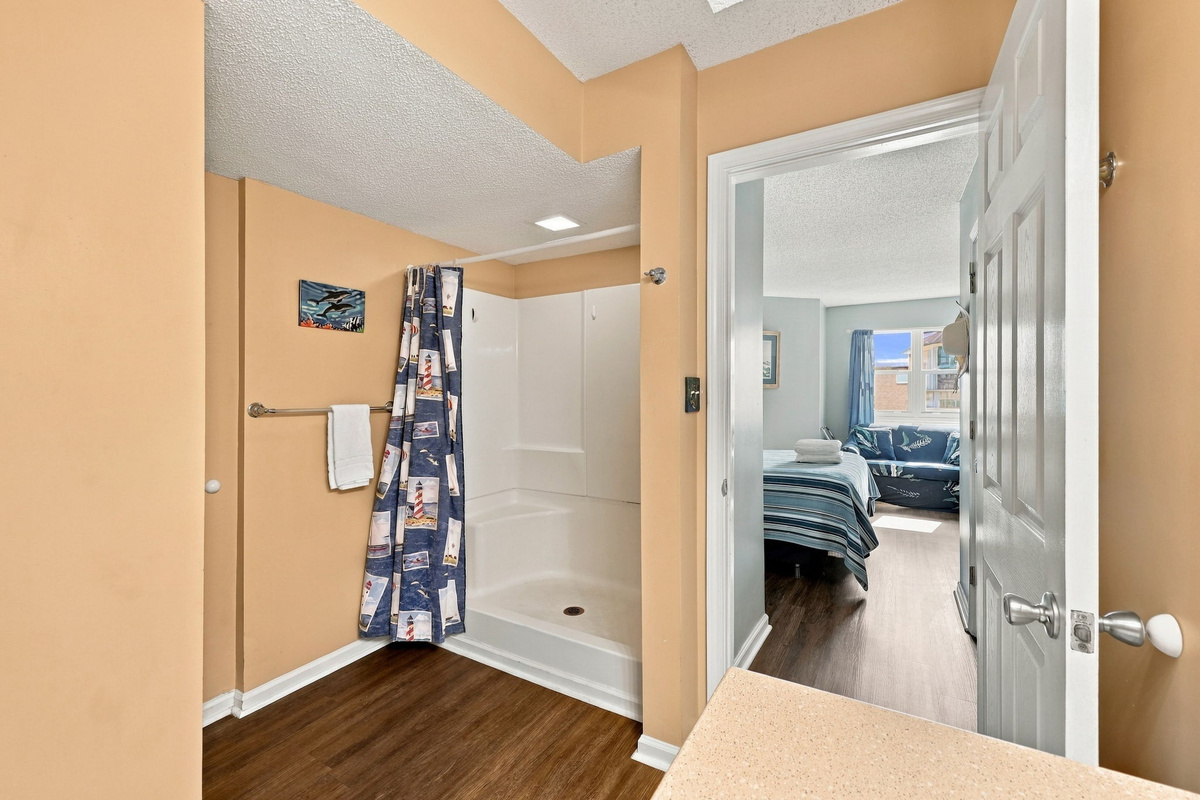 Ensuite master bathroom view highlighting warm beige walls, a walk-in shower with a coastal shower curtain, and an open doorway looking through to the light-filled master bedroom.
