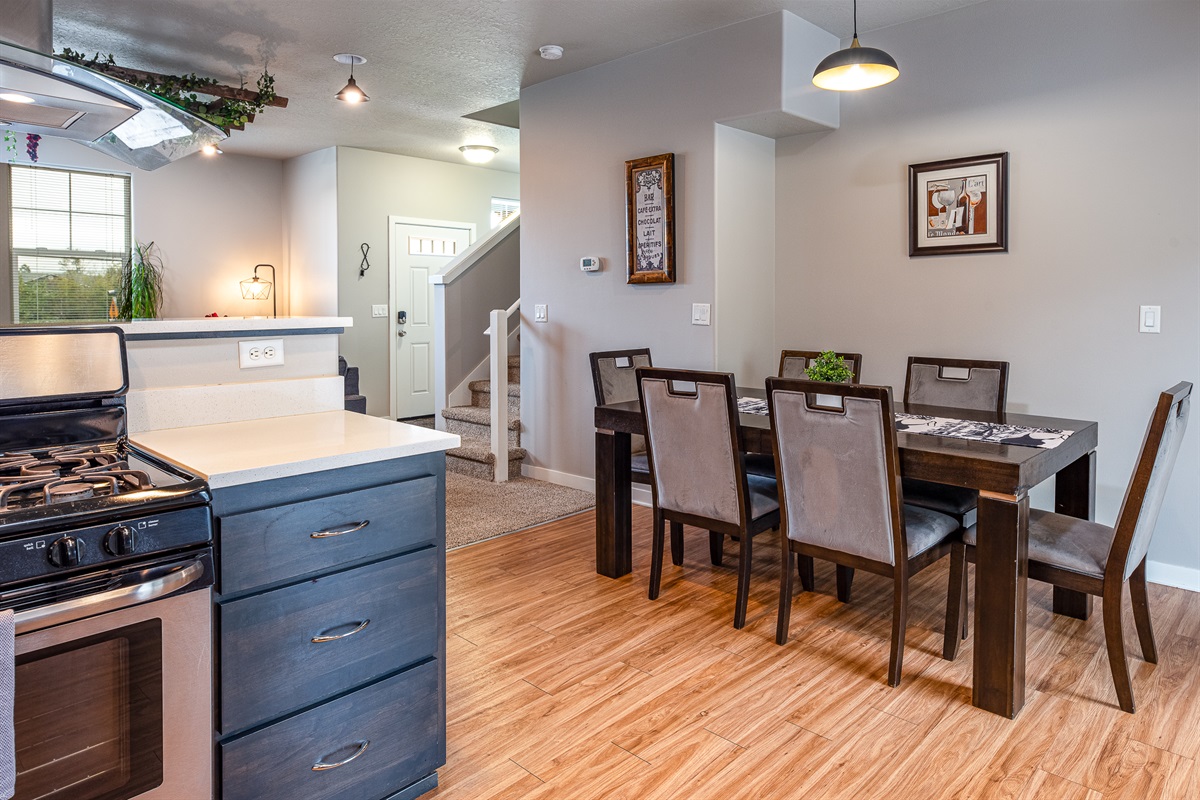 A modern kitchen and dining area with a black gas stove, light-colored countertops, a wooden dining table with chairs, and stairs leading to an upper level, complemented by decorative wall art and a door leading outside.