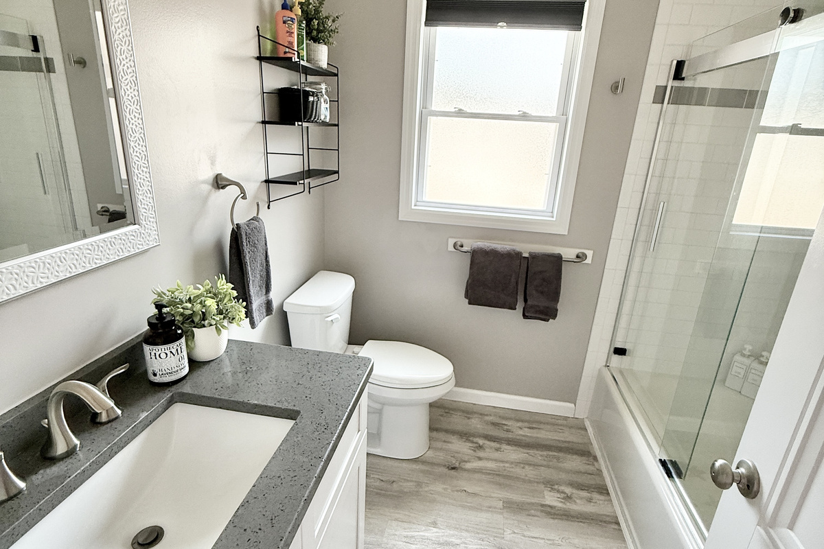 Refresh in this bright, updated bathroom featuring a sleek grey quartz vanity top and a white shaker-style cabinet. The space is finished with clean white walls, dark charcoal towels, and a window that provides plenty of natural light.