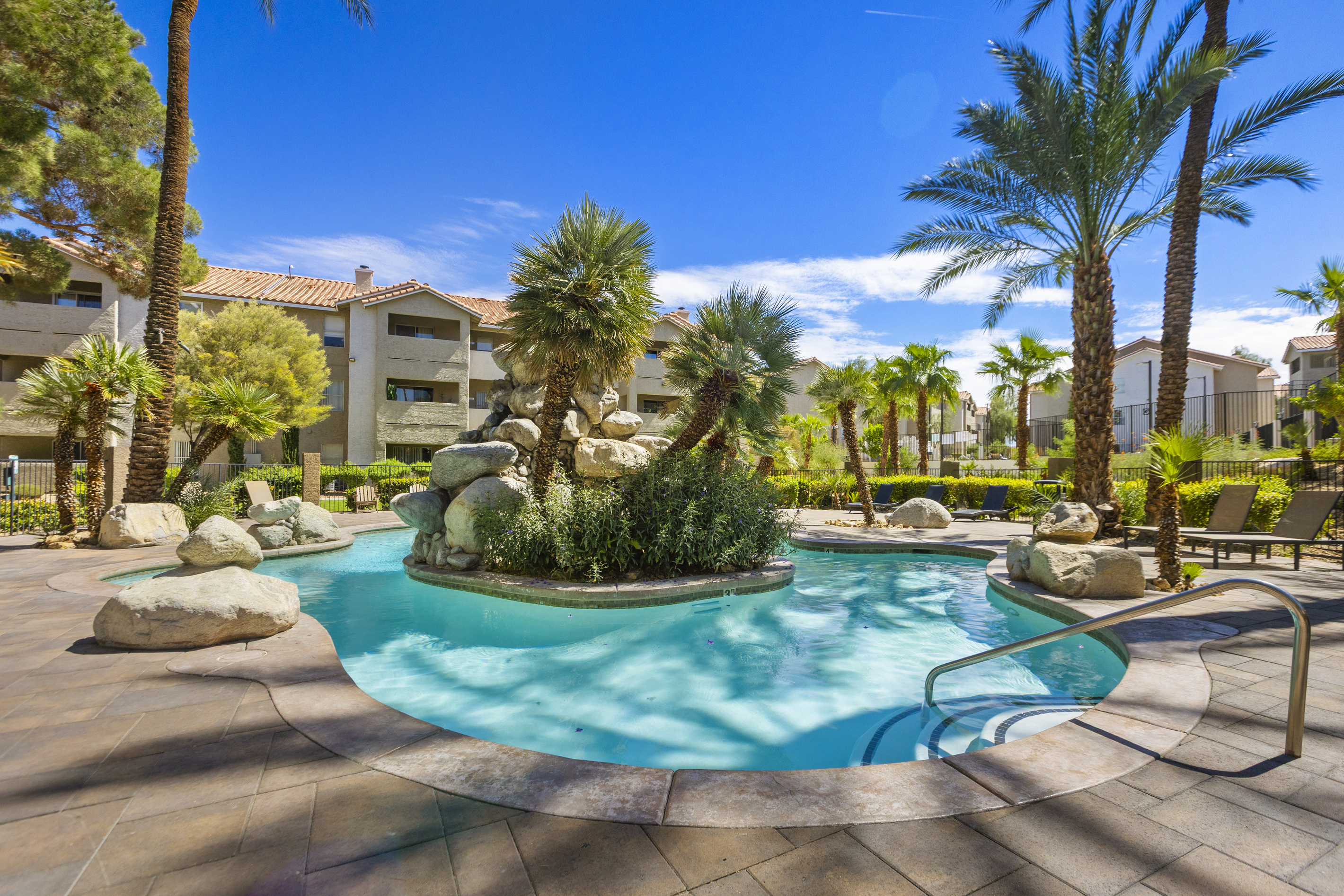 Shared resort-style pool 1 with rock waterfall, palm trees, paved lounge area, and shaded spots around the water.
