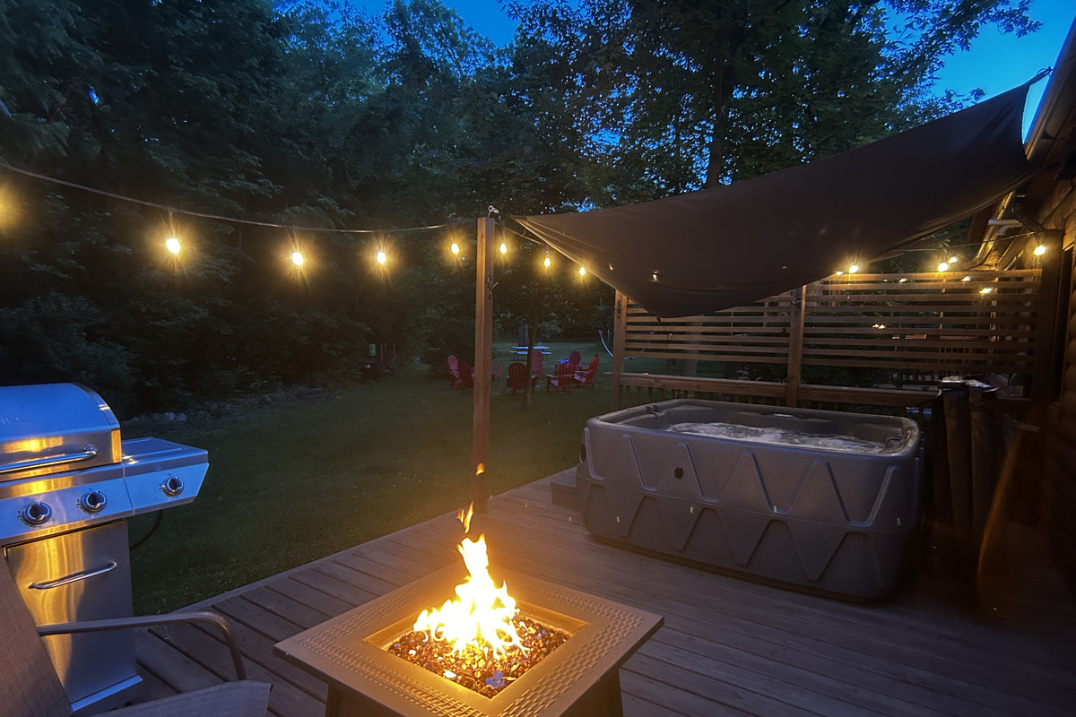 Private deck with fire table, seating, BBQ grill, hot tub, and glowing café lights—perfect for relaxing nights outdoors.