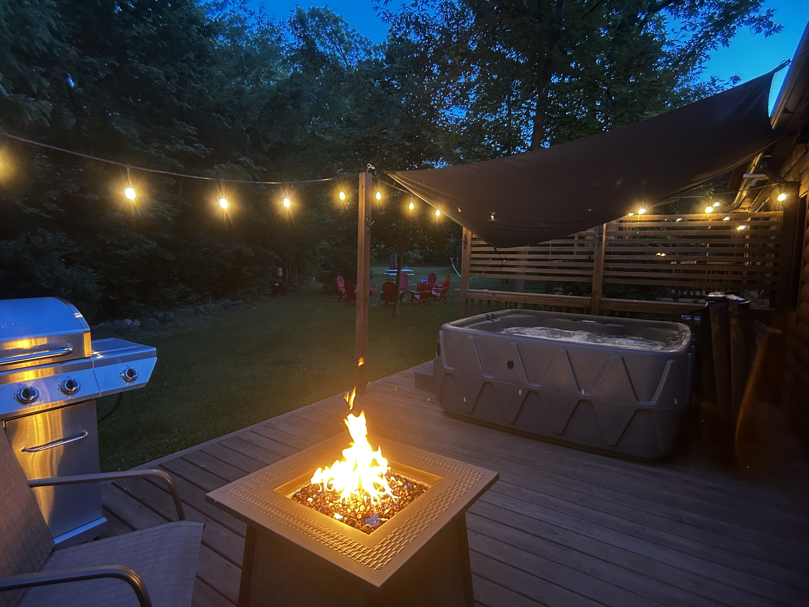 Private deck with fire table, seating, BBQ grill, hot tub, and glowing café lights—perfect for relaxing nights outdoors.