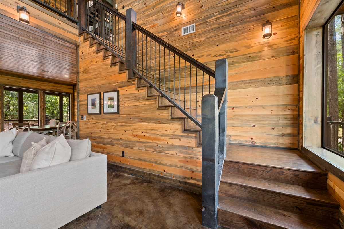 The living room features a modern staircase with black railings, leading up beside a wall of mixed wood planks, offering a glimpse into the open-plan space above.
