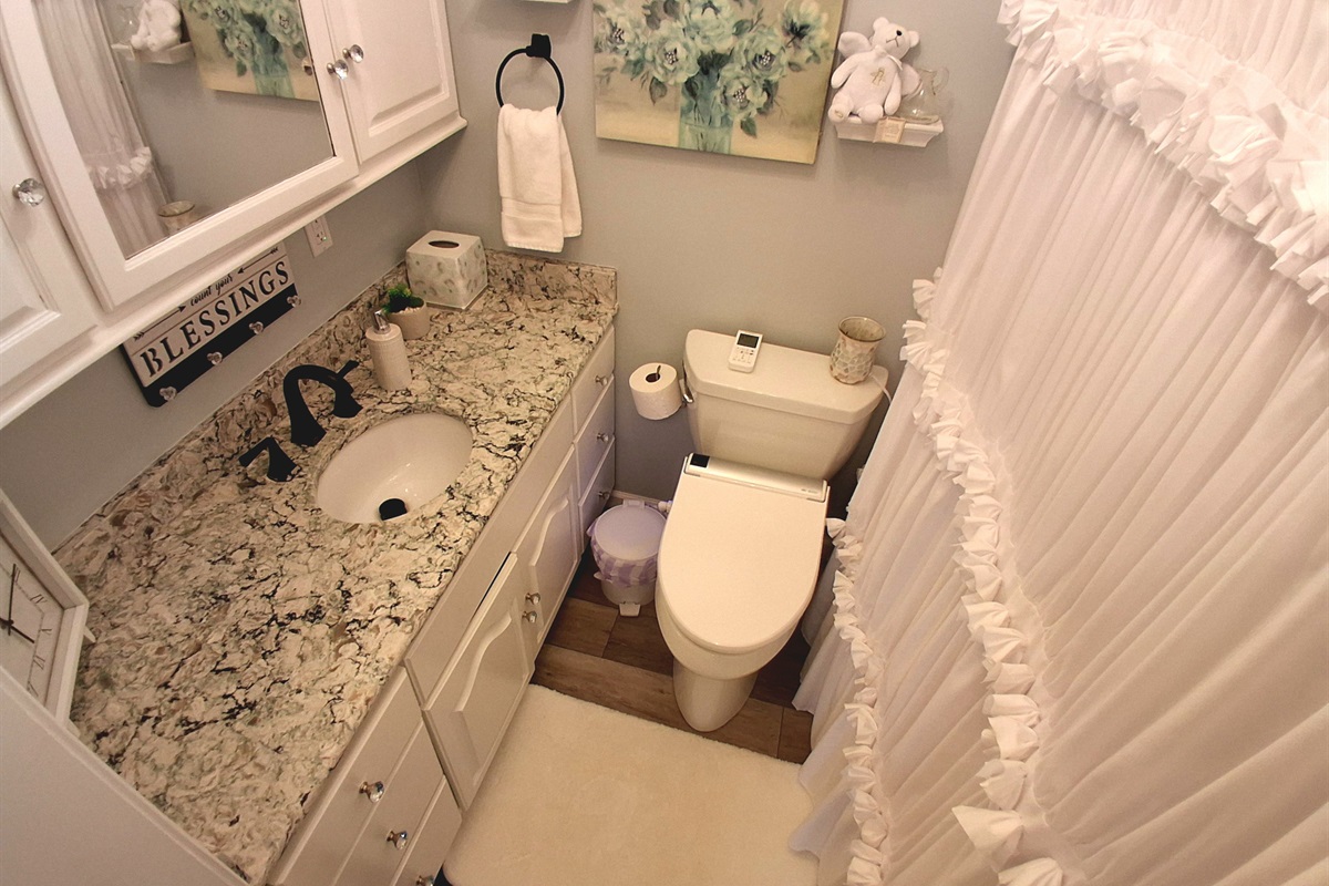 Pale blue walls offer a refined contrast to the crisp white décor, with a romantic ruffled shower curtain and vintage-style vanity. A sparkling marbled countertop adds beauty and space, creating a serene, elegantly styled sanctuary.