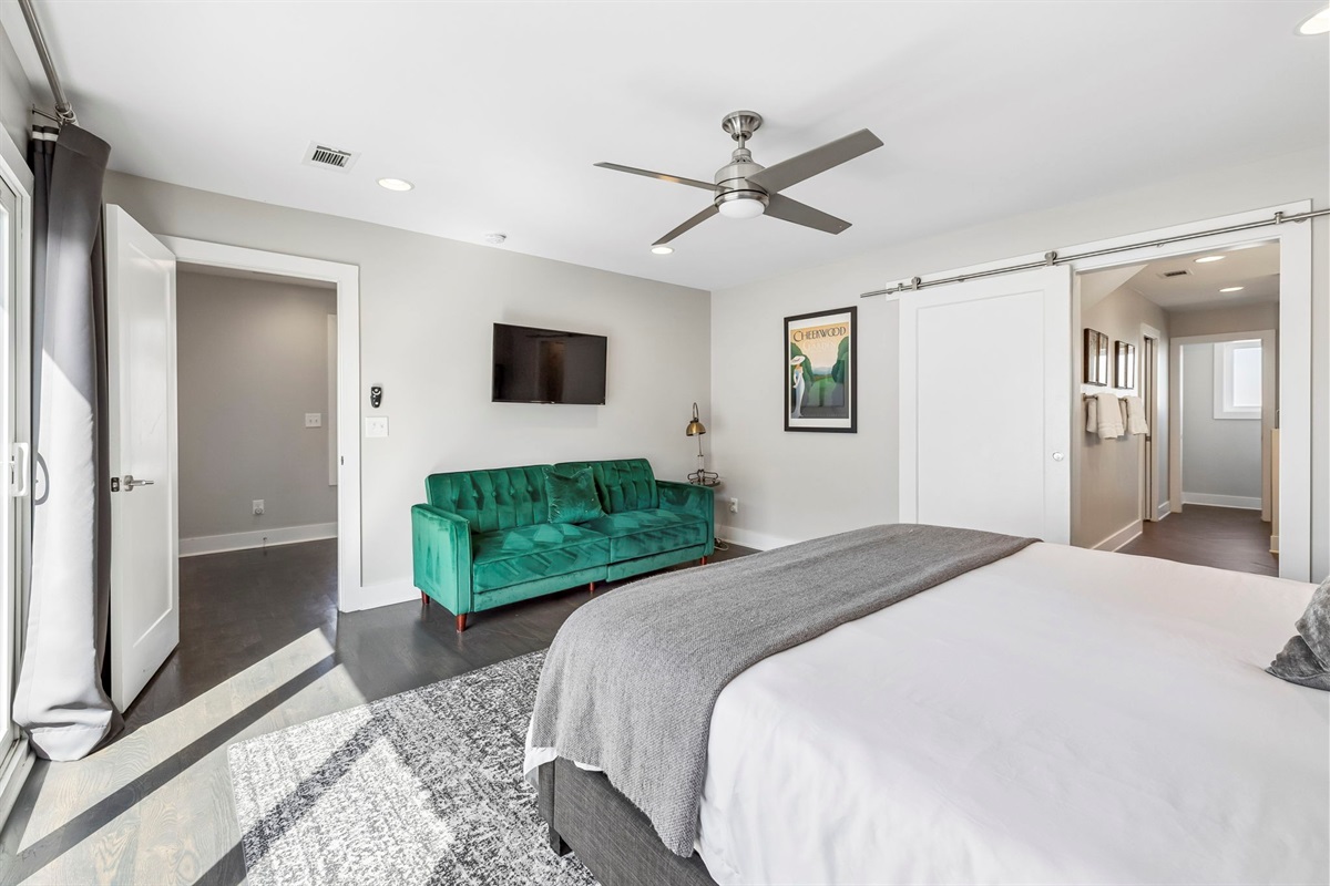 Emerald green tufted sofa adds a vibrant pop of color to this spacious bedroom lounge - Wall mounted TV and large windows make this a great spot for relaxing or streaming - Sophisticated black out drapes and detailed rug complete the room&rsquo;s upscale feel