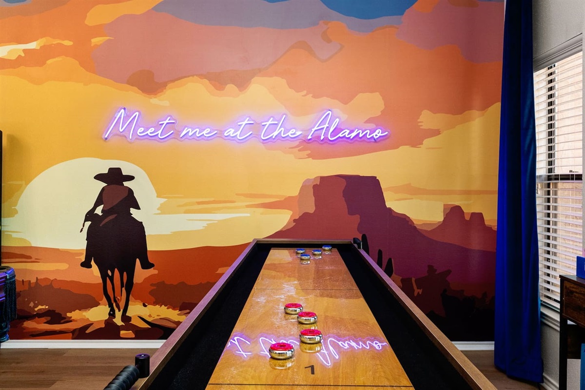 This vibrant Western-themed game room features a shuffleboard table set against a bold mural with a cowboy silhouette and neon “Meet me at the Alamo” sign—bringing Texas pride and playful energy to every game night.