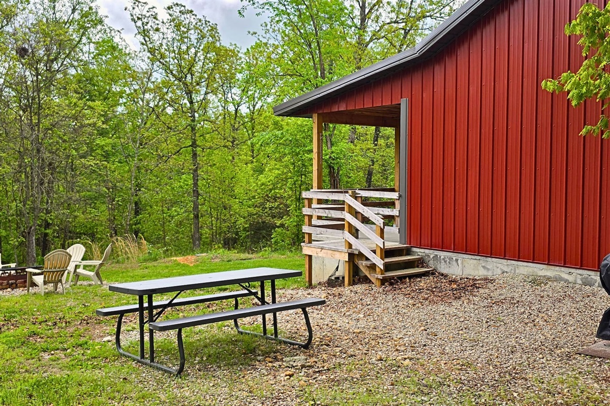 The backyard at Misty Oak Lodge invites you to unwind—featuring a charcoal grill, picnic table, and cozy firepit ringed with Adirondack chairs, all set against a peaceful wooded backdrop perfect for s’mores and starry nights.