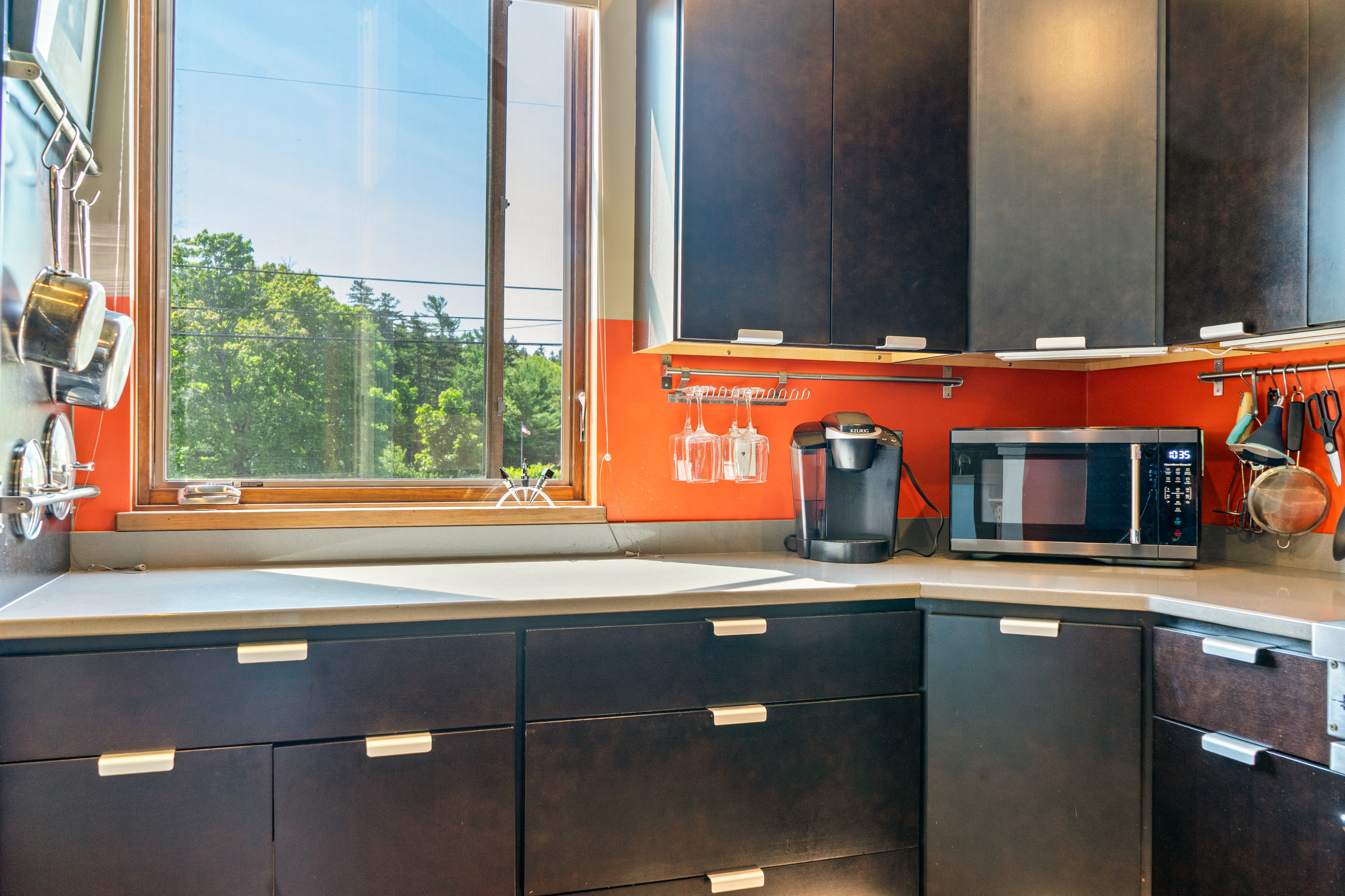 The Social Heart's gourmet kitchen nook features dark modern cabinetry, vibrant accents, a Keurig coffee station, and dedicated utensil racks. A sunlit window frame brings forest canopy views right to your prep space at "The Crag."