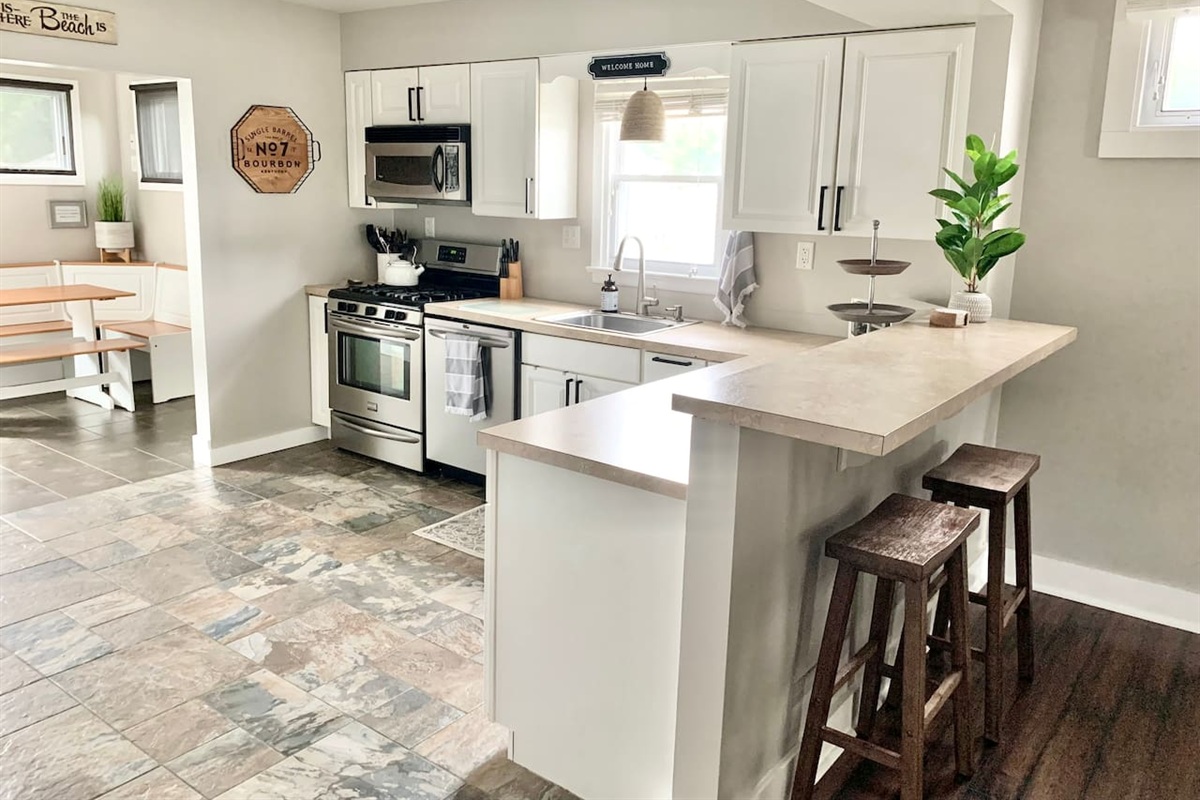 This modern kitchen features stainless steel appliances, a deep sink, and a raised breakfast bar with wooden stools. The open layout leads directly into a sunlit corner breakfast nook, providing a cozy spot for morning coffee.