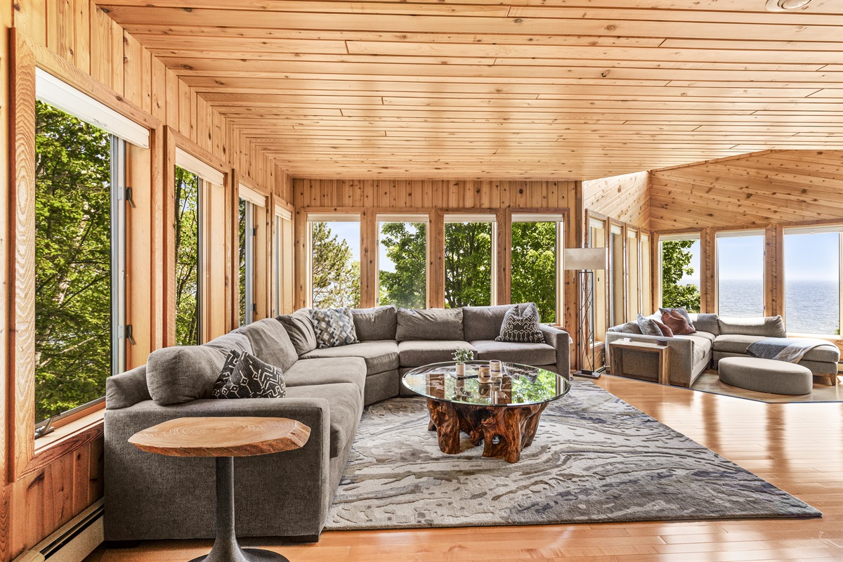 Spacious lakeview living room with a large comfortable sectional, stylish glass coffee table with a natural root-style base, and oversized windows showcasing Lake Superior, shoreline and surrounding trees. A beautiful gathering space designed for relaxing.