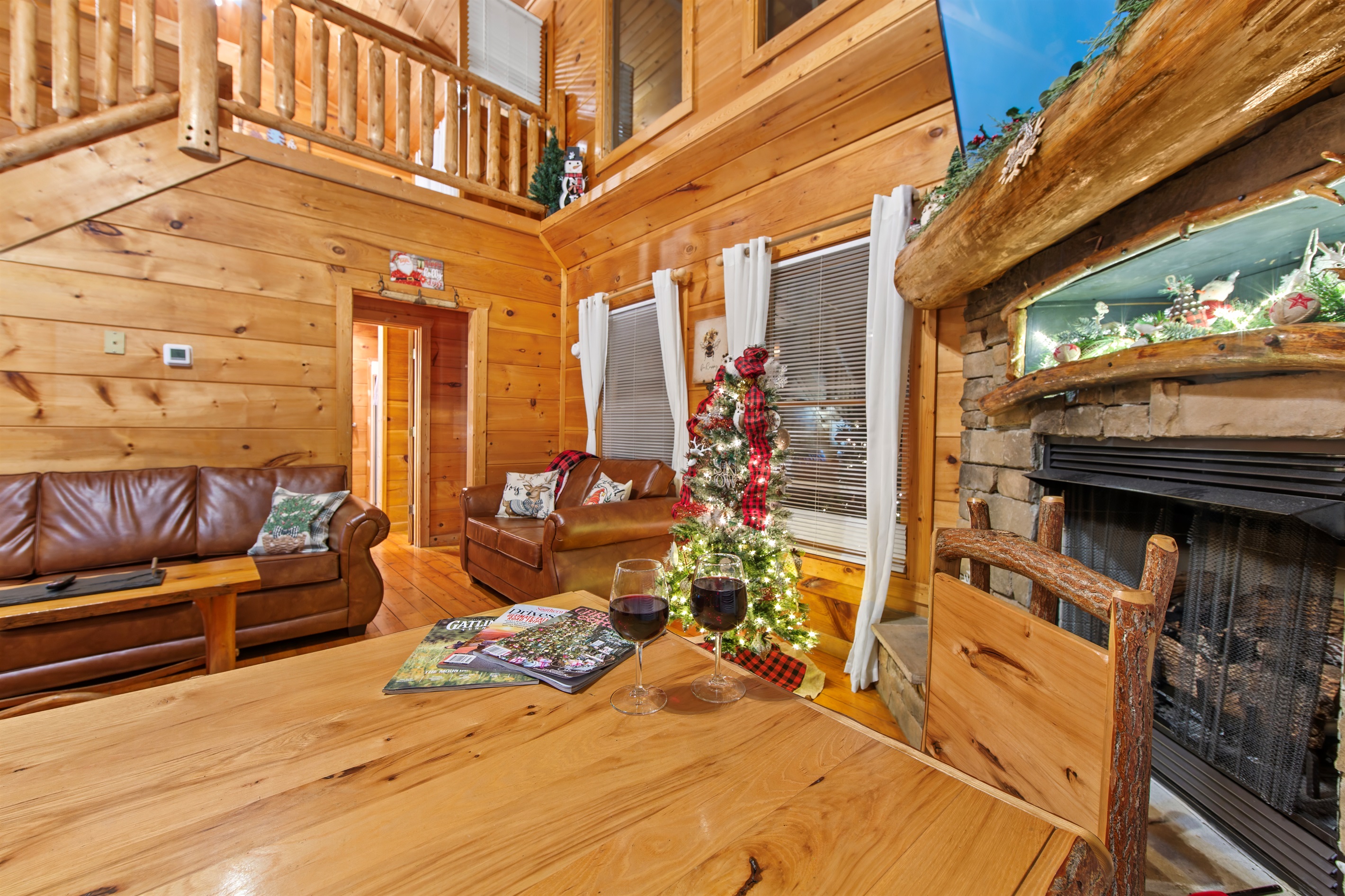 The cozy living room features warm wood paneling, a stone fireplace adorned with rustic decorations, and a leather couch with pillows, creating a welcoming cabin retreat.