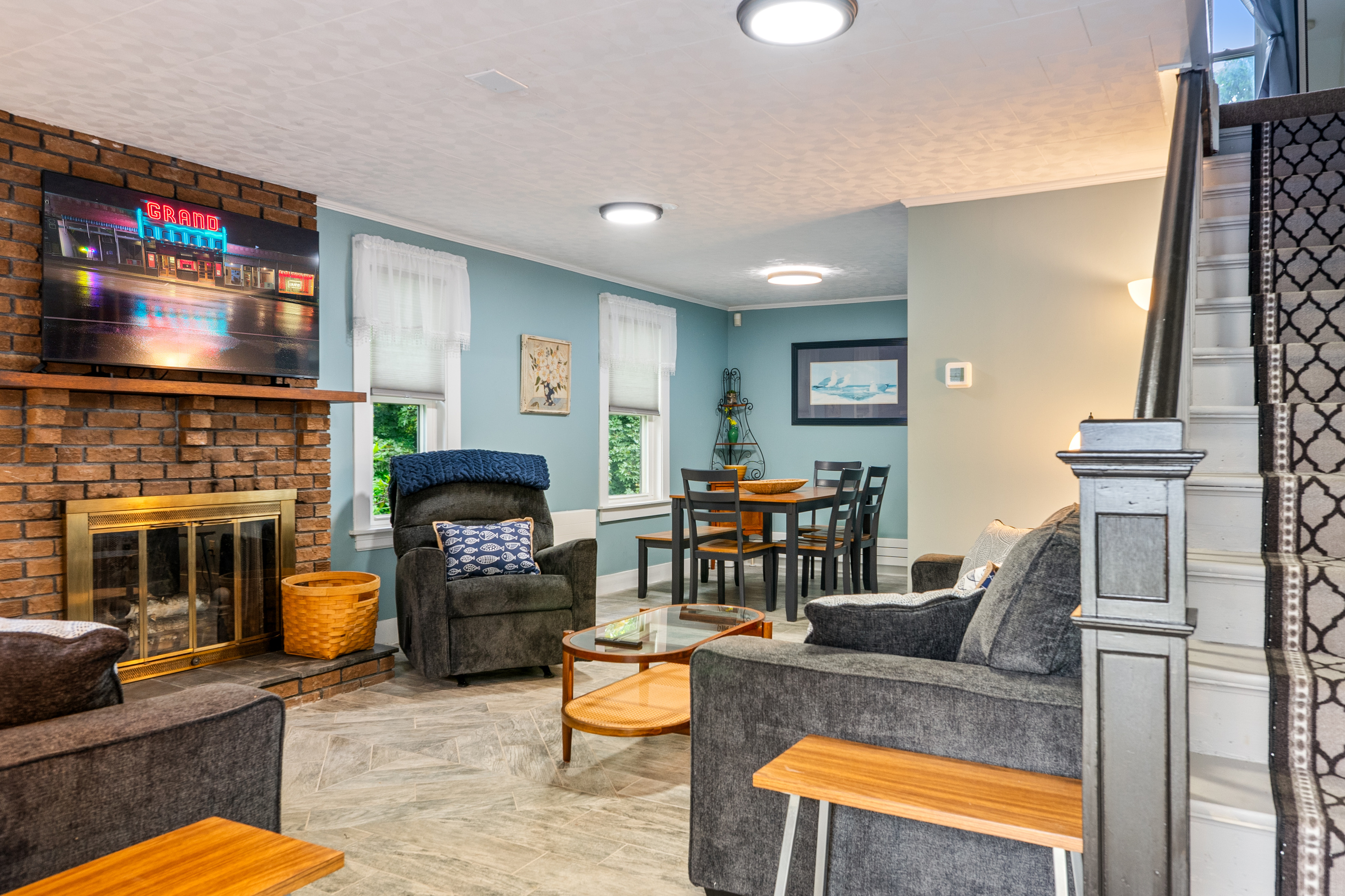 A smart TV sits on the mantel of the mid-century brick fireplace, a prominent design feature in the living room. The open-concept layout reveals the adjacent dining table and second-story staircase, mapping out the home’s intuitive dual-level flow.
