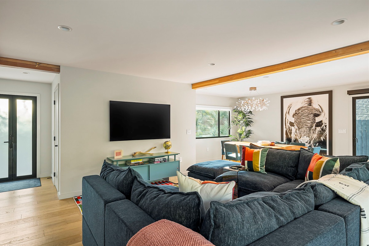 A modern open-concept living area featuring a large sectional sofa, a wall-mounted television, and exposed wooden beams, with a dining area and a large framed buffalo print visible in the background.